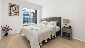 Ein modernes Schlafzimmer mit einem großen Bett, einem Nachttisch mit Lampe und Blick auf Gebäude durch das Fenster.