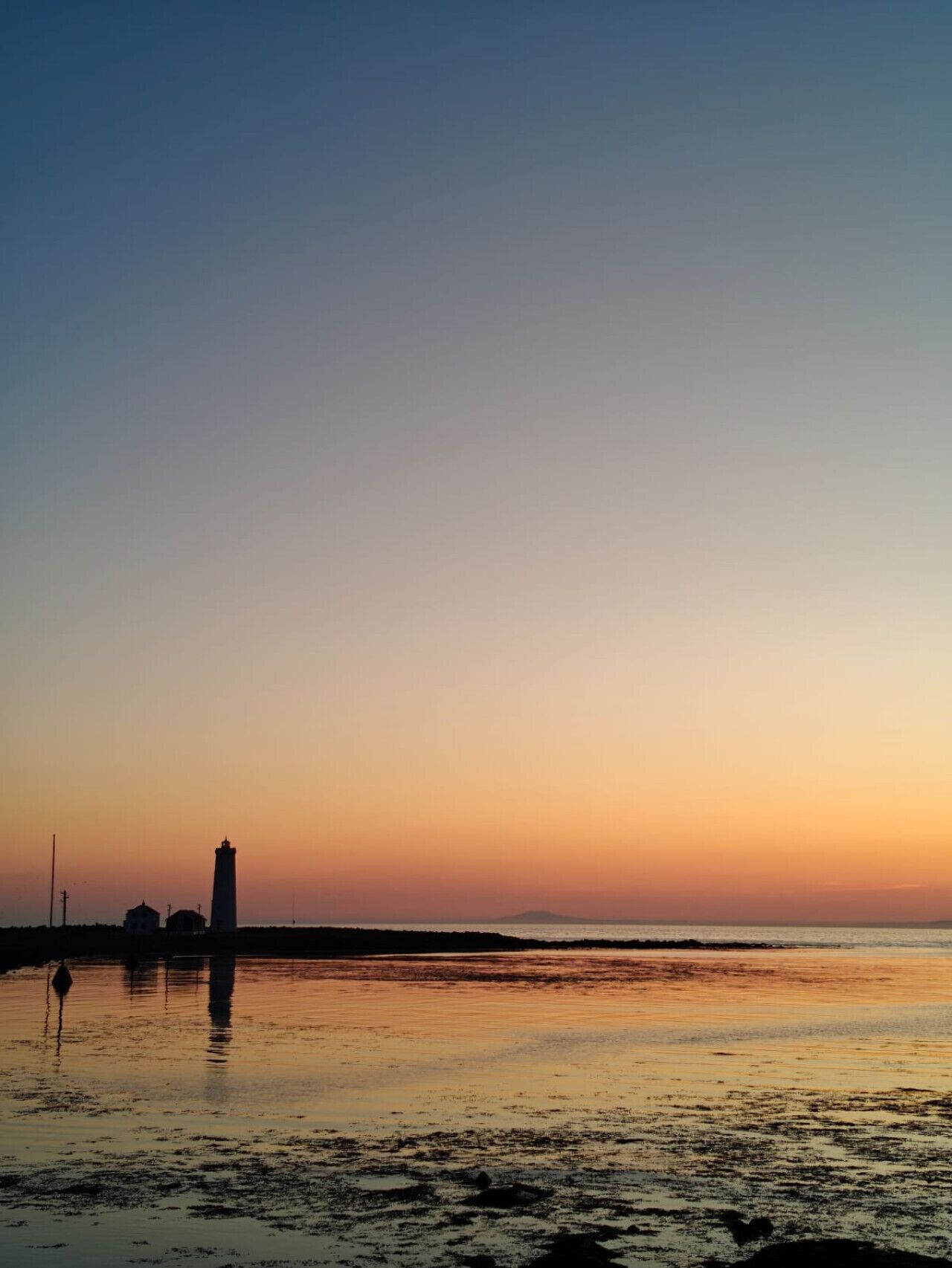 A coastal scene with a lighthouse and buildings silhouetted against a sunset sky.