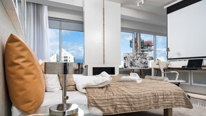 Habitación de hotel moderna con una cama grande, una mesa de centro de vidrio, un escritorio con una computadora portátil y un ventanal que ofrece vistas a la ciudad.
