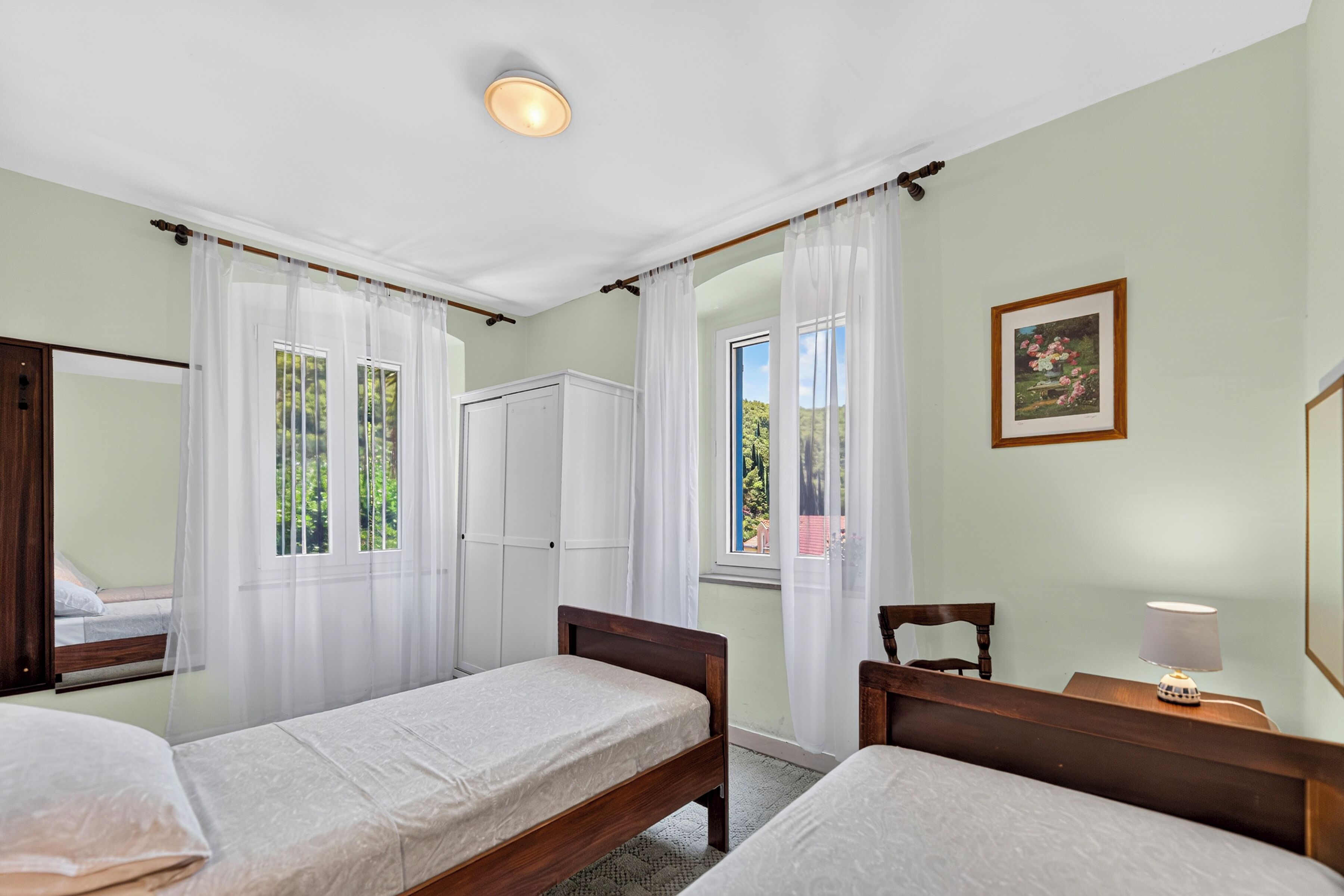 A bedroom with two single beds, a wardrobe, a bedside table with a lamp, and a window with a view of greenery.