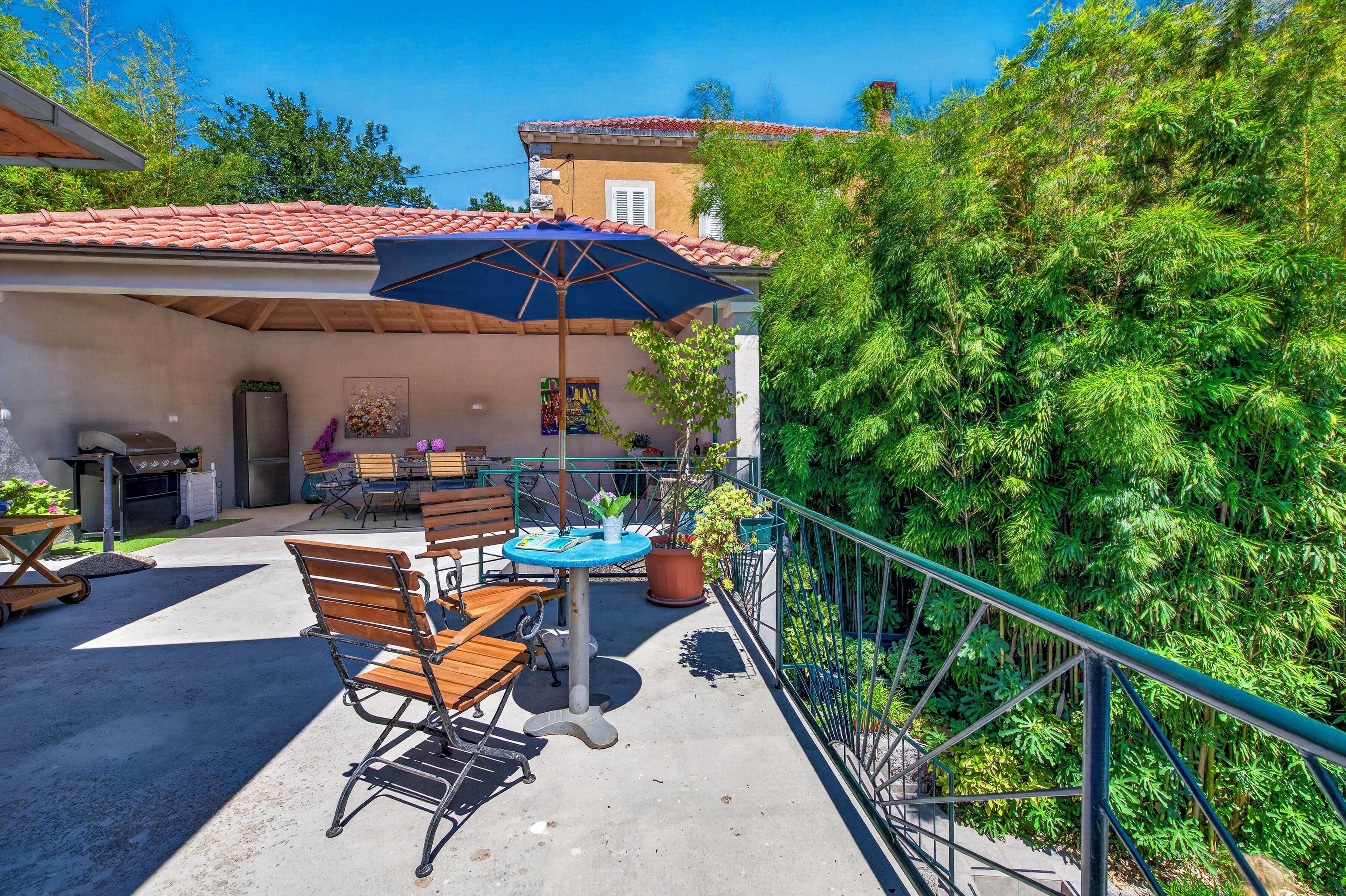 Un espace patio aménagé avec une table et des chaises, un parasol bleu et une balustrade donnant sur un jardin.