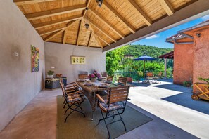 Un espace extérieur aménagé pour les repas, avec une table et des chaises en bois, entouré d’une terrasse couverte d’où l’on peut admirer une colline verdoyante.