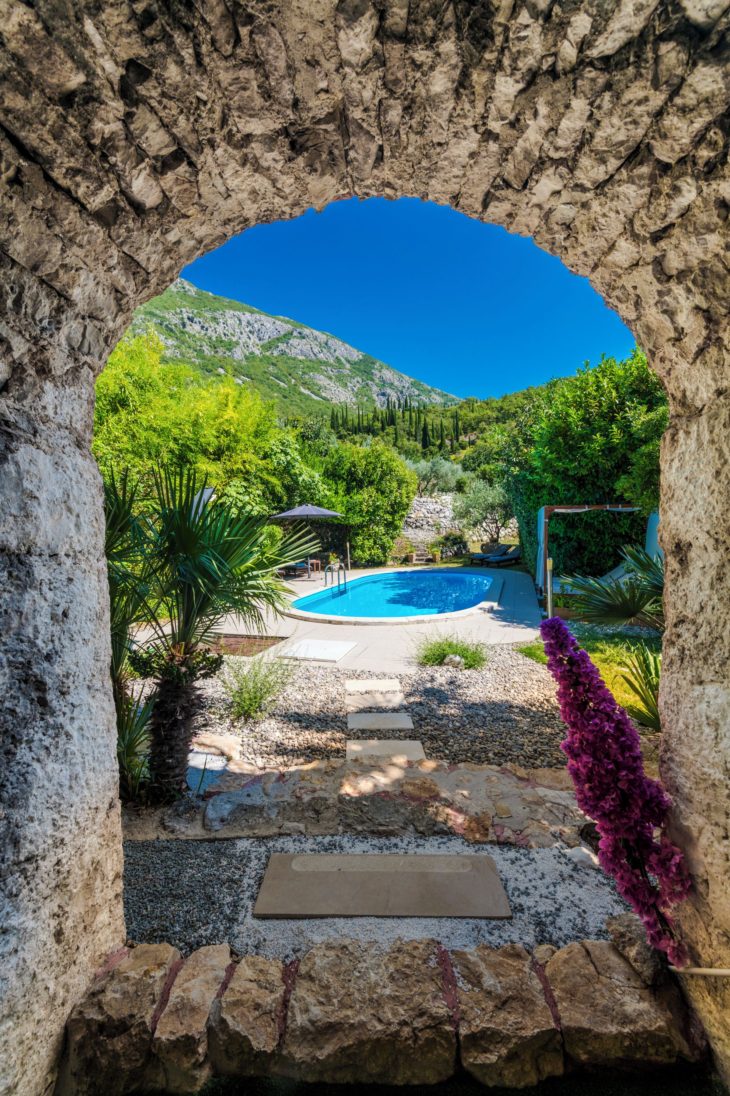 Un arco de piedra que da acceso a una zona de piscina con montañas al fondo.
