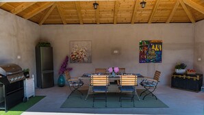 Eine Außenpatio mit Holzboden, Grill, Kühlschrank, Esstisch mit Stühlen und Kunstwerken an den Wänden.
