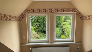 A bedroom with a sloped ceiling, two windows with a view of greenery, a radiator, and a bed with a textured blanket.