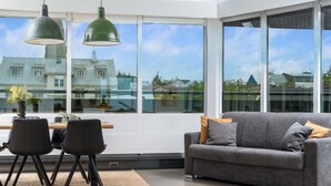 Ein modernes Wohnzimmer mit einem grauen Sofa, zwei schwarzen Sesseln und einem Tisch mit Topfpflanzen. Große Fenster bieten Blick auf Gebäude und Bäume.