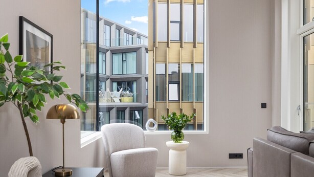 Un salon moderne donnant sur un immeuble grâce à de grandes fenêtres, avec un canapé gris, un fauteuil blanc, une petite table avec une lampe et une plante en pot.
