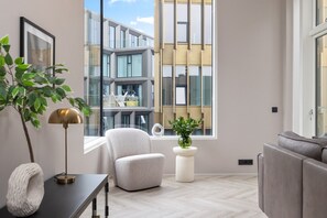 A modern living room with a view of a building through large windows, a grey sofa, a white armchair, a small table with a lamp, and a potted plant. - Most Desirable Penthouse in (Reykjavík)