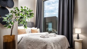 Une chambre d’h ôtel moderne avec un grand lit, une plante en pot, une petite table ronde avec une lampe, et une vue sur la ville par la fenêtre.