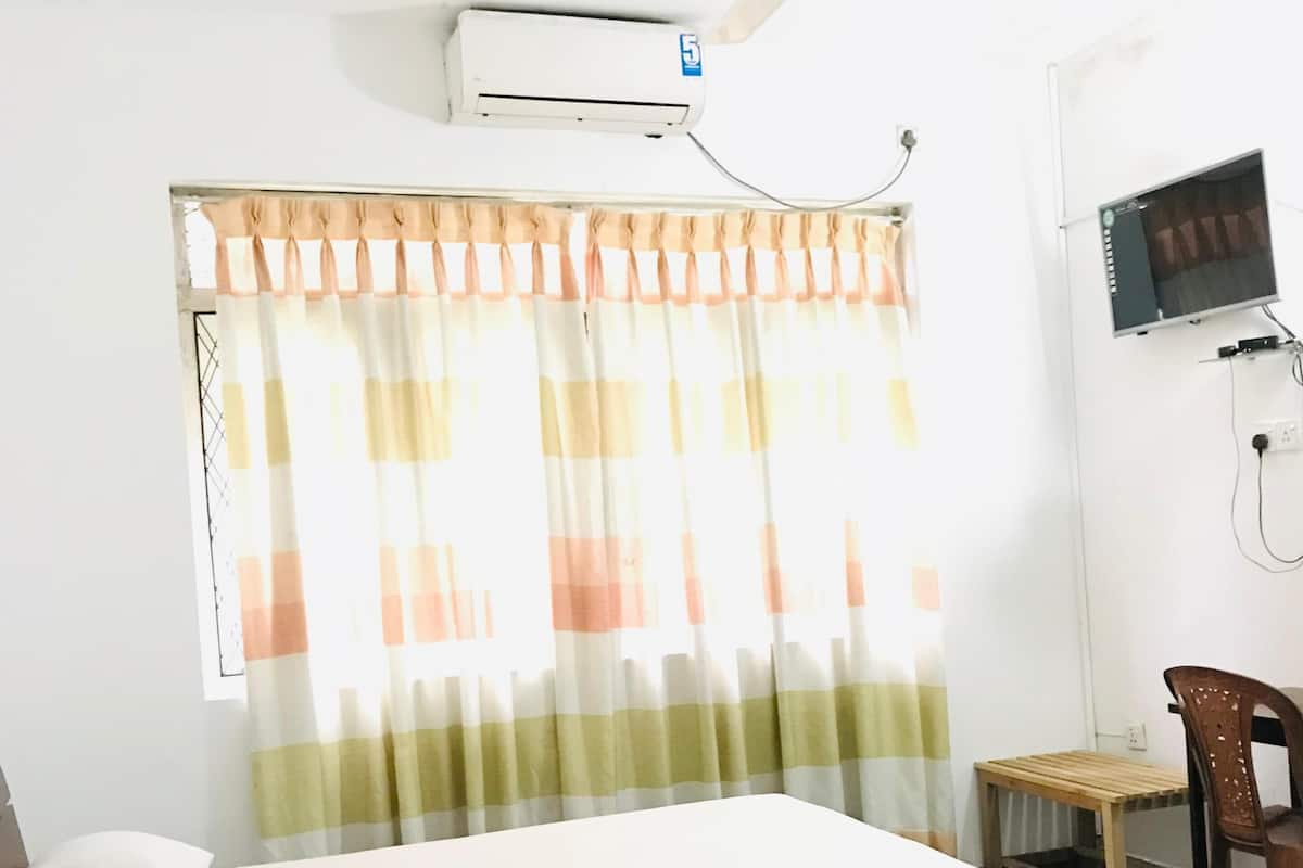 A bedroom with a bed, a ceiling fan, an air conditioning unit, a TV, and a window with curtains.