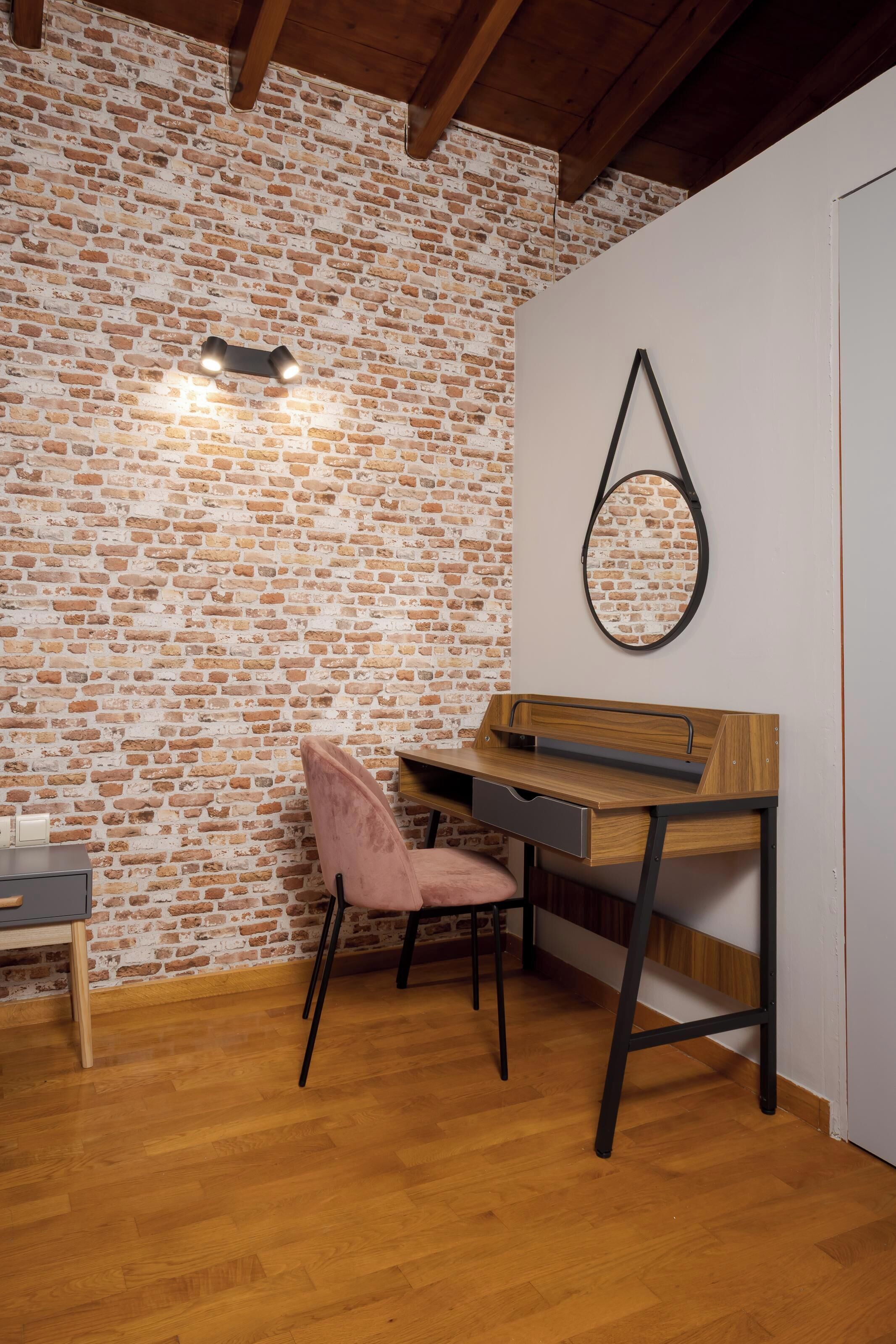 A room with a brick wall, a wooden desk, a pink chair, and a mirror on the wall.