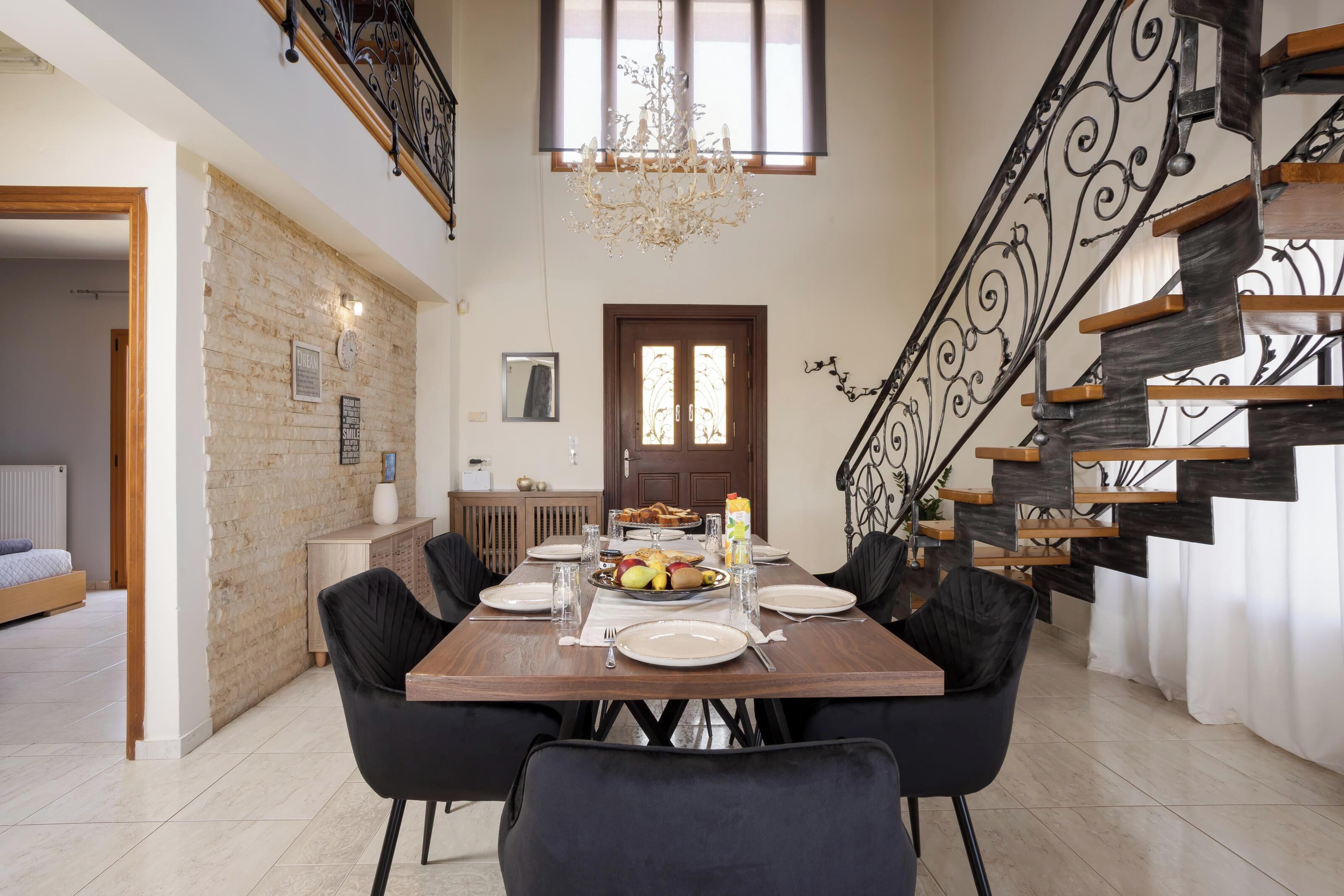 A dining area with a wooden table set for four, surrounded by black chairs. A chandelier hangs from the ceiling, and a staircase with wooden steps and metal railings is visible to the right.