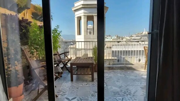 A balcony with a view of a tower and buildings. There are wooden chairs and a table. - Comfy penthouse in the center of Athens (Athina)