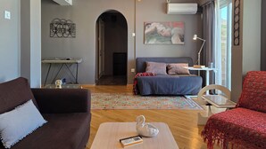 A modern living room with a sofa, a coffee table, and a bed. There is a desk with a computer and a lamp. The room has a large window and an air conditioning unit on the wall. - Comfy penthouse in the center of Athens (Athina)