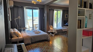 A hotel room with a bed, a desk, a chair, a bookshelf, and a view of the city through large windows. - Comfy penthouse in the center of Athens (Athina)