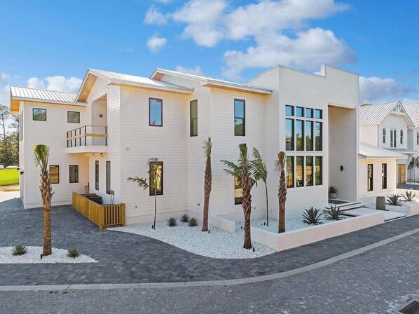 A modern two-story house with a flat roof, large windows, and a landscaped front yard with palm trees and shrubs.