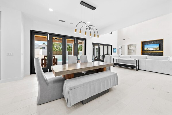 A modern living room with a large dining table, grey sofas, and a TV mounted on the wall. - 5-bedroom house in Saltwater, Walk to Beach, Private Pool, Bikes, Furry Friends (Santa Rosa Beach)