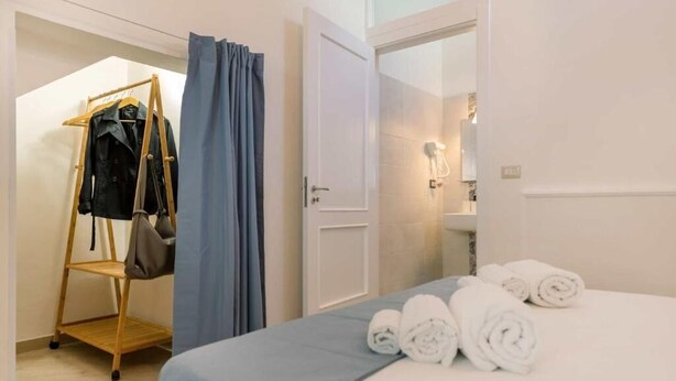 Ein Hotelzimmer mit einem Bett, einem hölzernen Kleiderhänger mit Kleidung, einem Badezimmer mit Spiegel und einem Handtuchhalter.