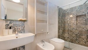 Ein Badezimmer mit einem weißen Waschbecken, einem Bidet, einer Toilette, einer Duschkabine aus Glas und Fliesen mit Muster.