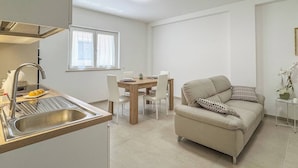 A modern kitchen with a built-in sink, dining area with a wooden table and chairs, and a beige sofa with patterned cushions.