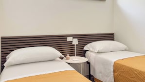 A hotel room with two beds, a small round table with a lamp, and a window with a view of the sky.