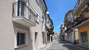 A narrow street with buildings on both sides, featuring balconies with railings and some with flower boxes.