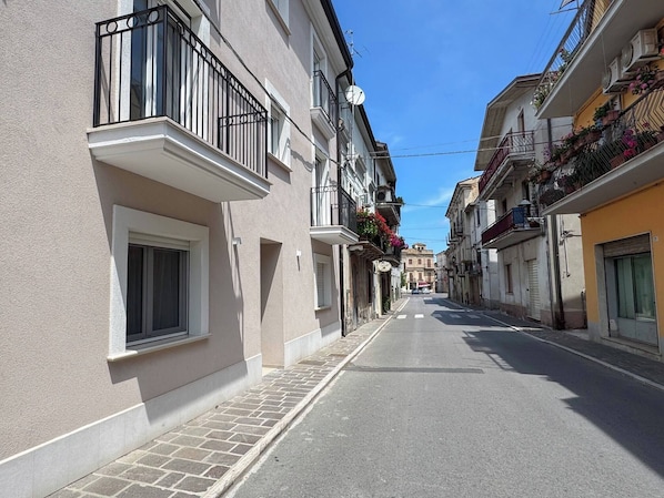 Une rue étroite bordée de bâtiments des deux côtés, avec des balcons dotés de garde-corps et certains agrémentés de jardinières.