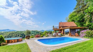 A swimming pool with a wooden deck and lounge chairs, surrounded by lush greenery and a view of mountains. - Trakoscan Castle View Pool House /w Private Spa (Bednja)