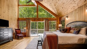 Une chambre spacieuse avec un grand lit, des murs en bois, un ventilateur de plafond et de grandes fenêtres donnant sur la forêt.