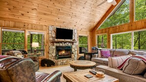 L’intérieur chaleureux d’un chalet avec une cheminée en pierre, des meubles en cuir et de grandes fenêtres donnant sur la forêt.