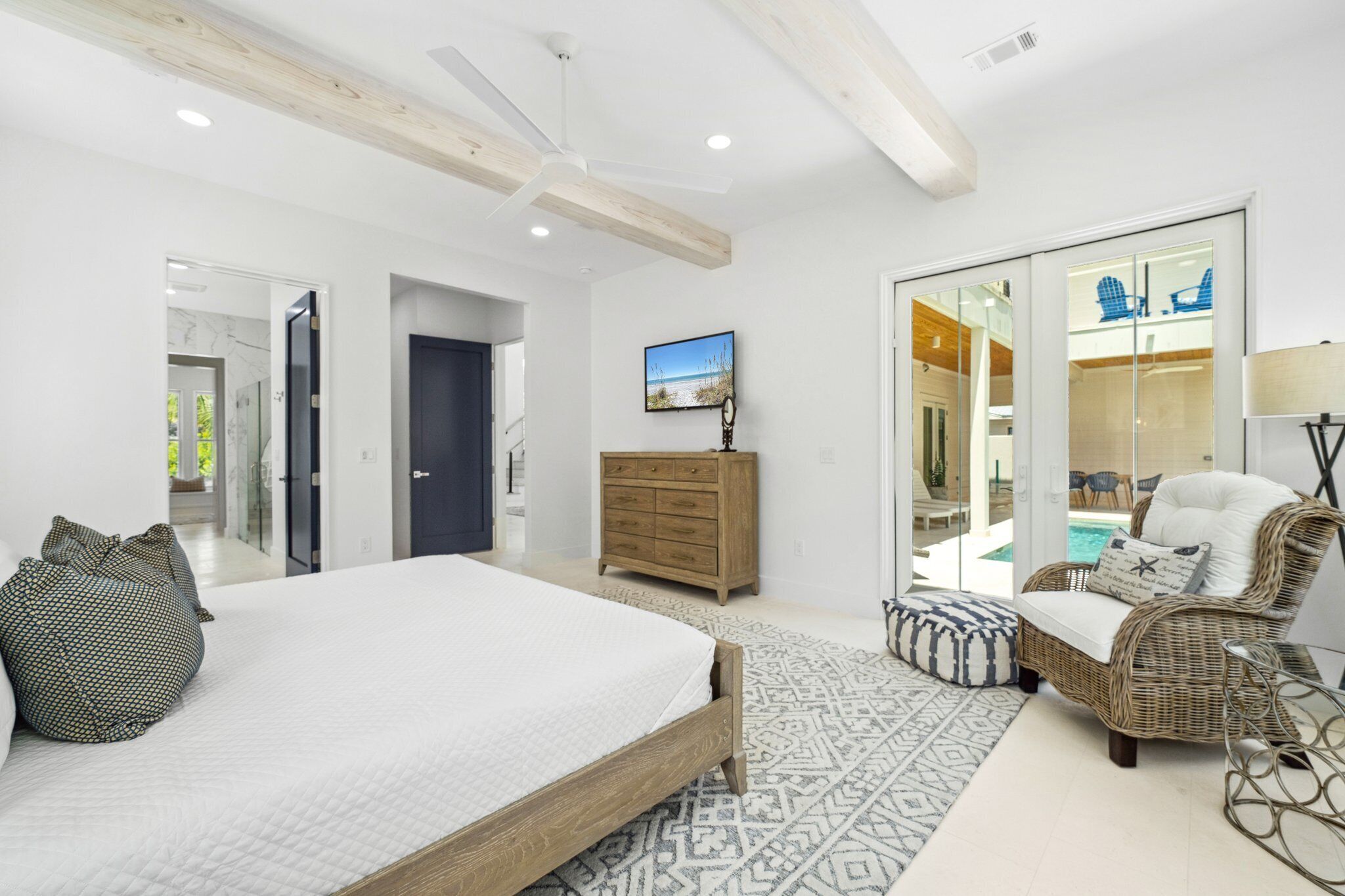 Een slaapkamer met een groot bed, een dressoir, een rieten stoel en een glazen deur naar een zwembad.