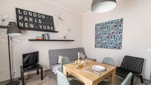 A modern living room with a wooden dining table, two chairs, a sofa, a TV, a floor lamp, a bookshelf, and a wall-mounted light fixture. - GuestHost - Lovely Apartment in Barletta (Barletta)