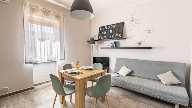 A dining area with a wooden table and chairs, a sofa, a TV, and a wall with a sign listing cities. - GuestHost - Lovely Apartment in Barletta (Barletta)