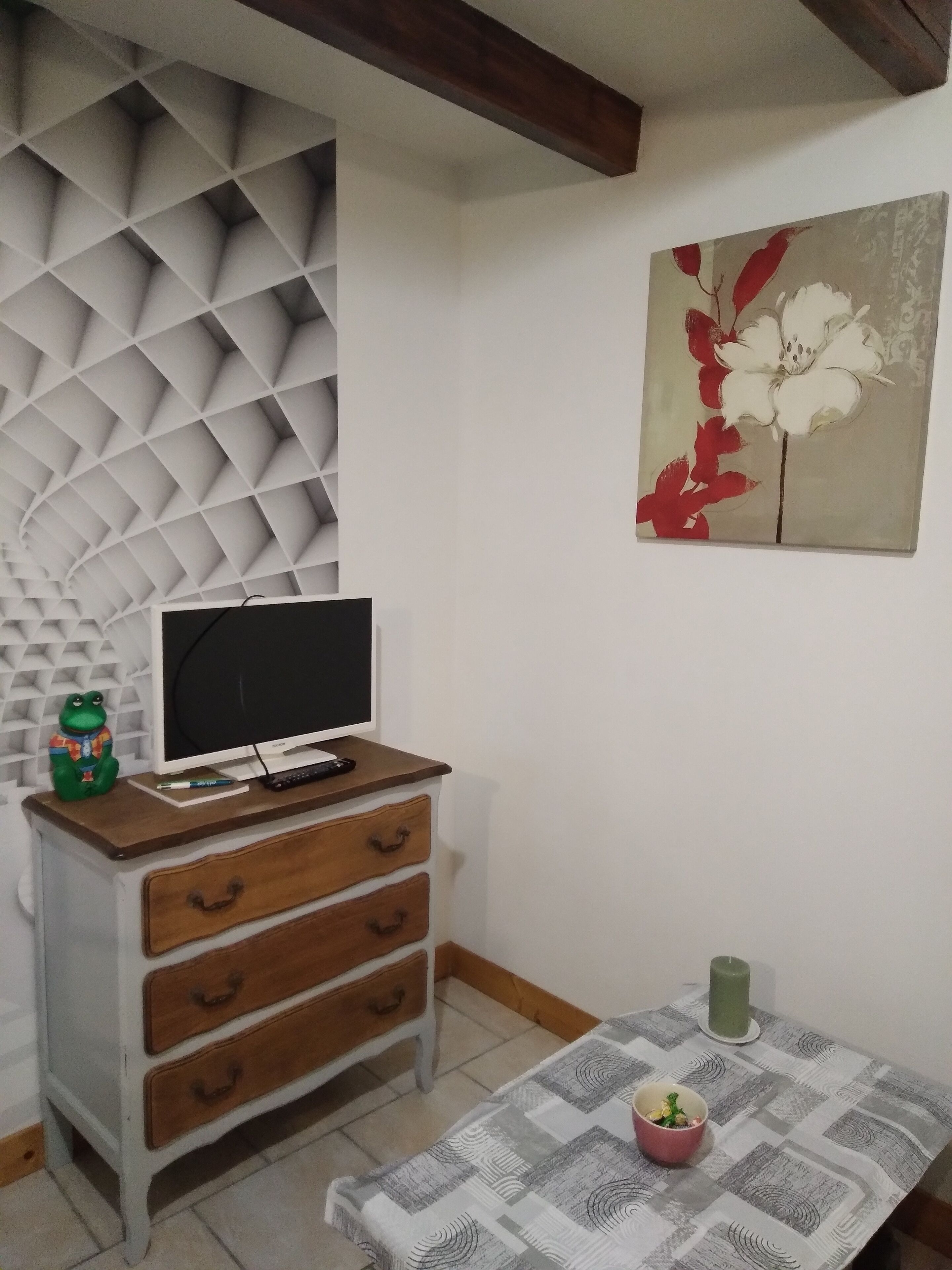 Ein Zimmer mit Fernseher auf einer Holzkommode, einer grünen Froschfigur, einem Blumenbild und einem Tisch mit gemustertem Tischtuch, sowie einer Kerze.