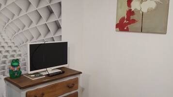 Ein Zimmer mit Fernseher auf einer Holzkommode, einer grünen Froschfigur, einem Blumenbild und einem Tisch mit gemustertem Tischtuch, sowie einer Kerze.