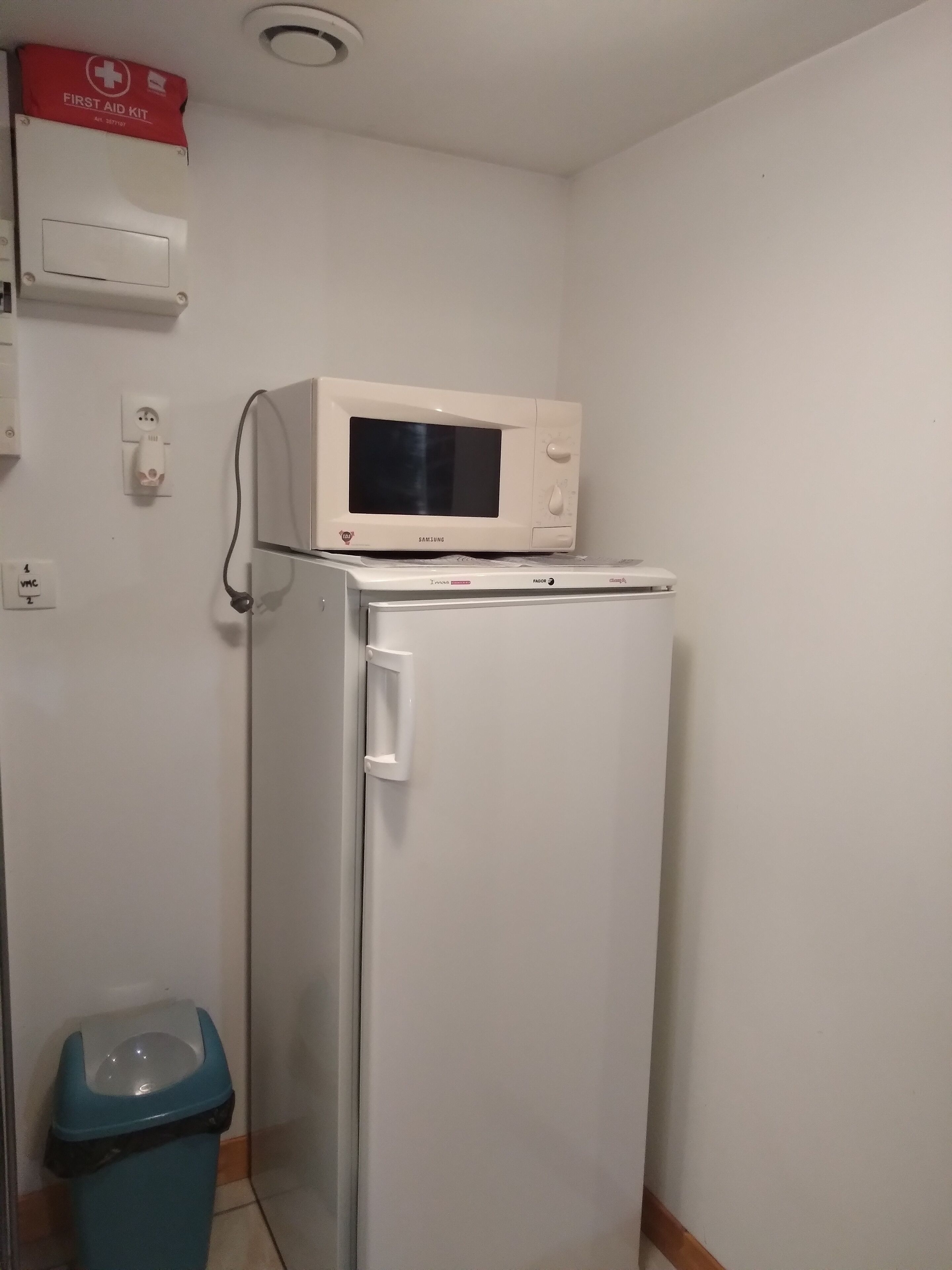A compact kitchen with a white refrigerator, a microwave on top, a trash bin, and a first aid kit on the wall.