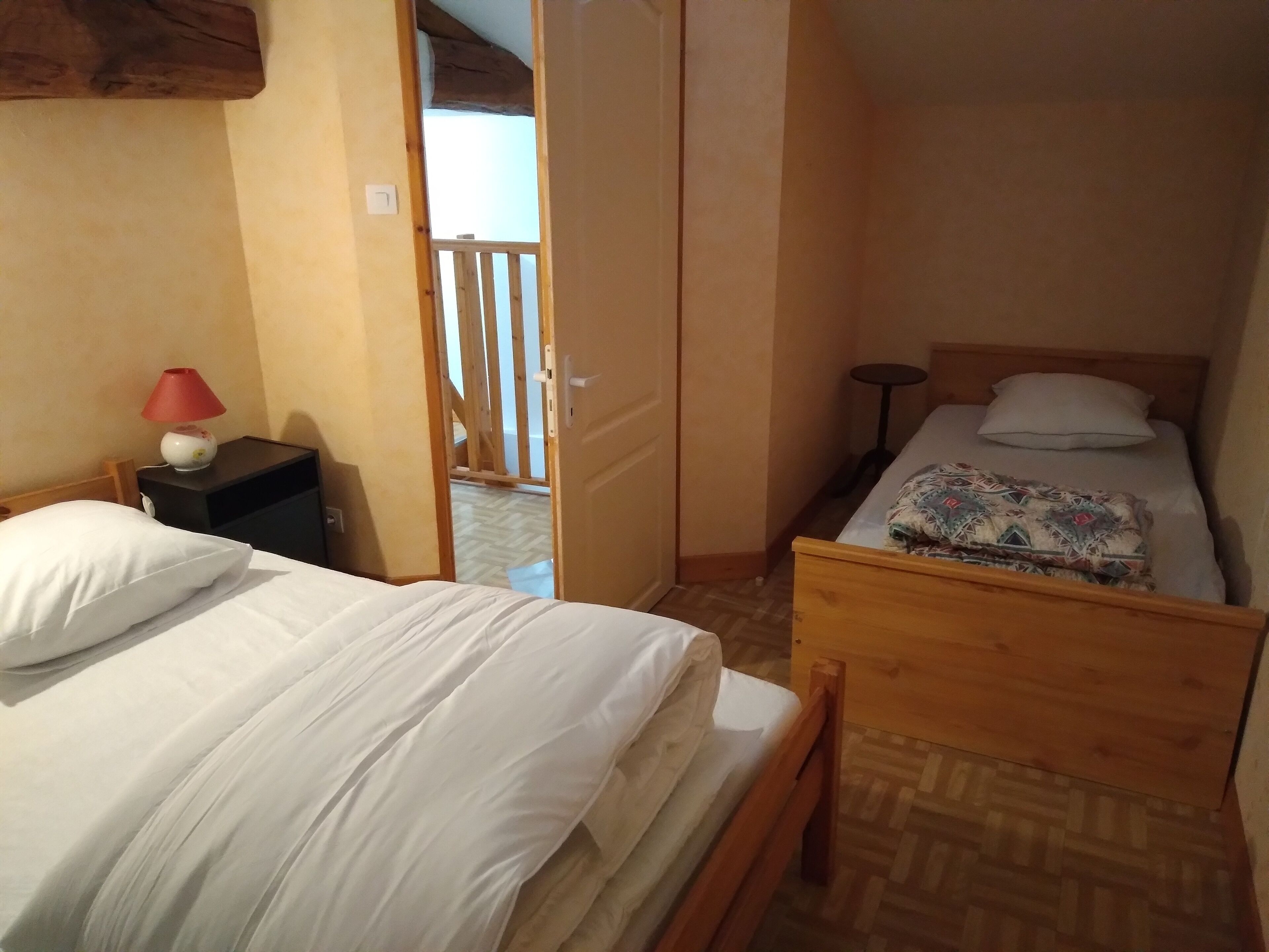 Um quarto pequeno e acolhedor com duas camas, uma mesa de cabeceira com abajur e uma porta de madeira que dá para área externa.