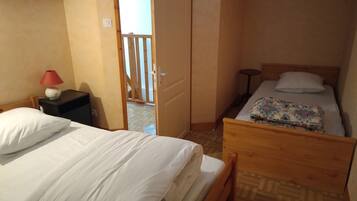 Um quarto pequeno e acolhedor com duas camas, uma mesa de cabeceira com abajur e uma porta de madeira que dá para área externa.