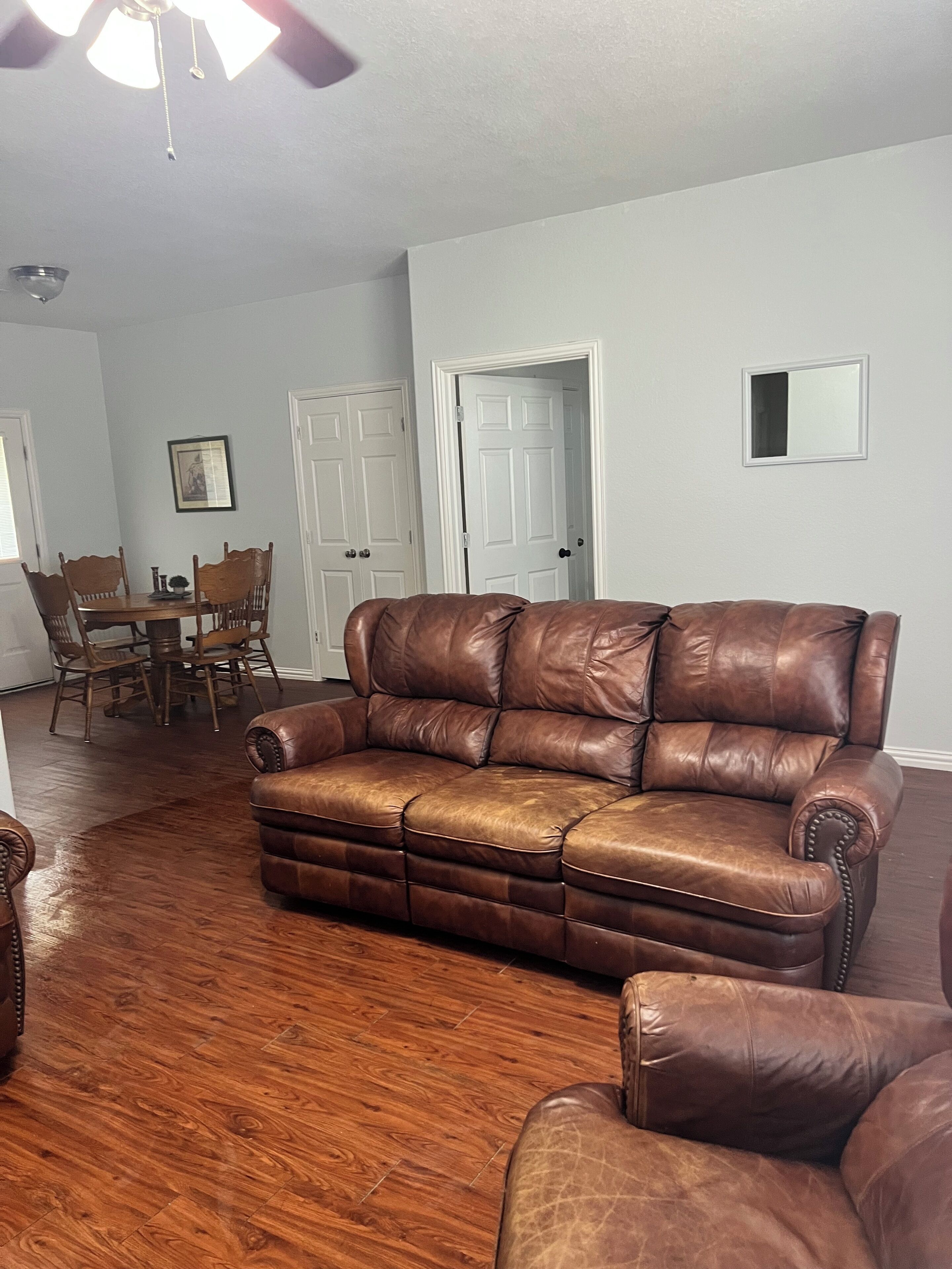 A living room with a brown leather sofa, wooden dining table, chairs, and a ceiling fan.