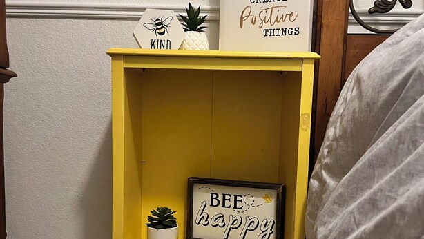 Una mesita de noche amarilla con una pequeña planta en maceta y un cartel enmarcado que dice 'BEE happy'.