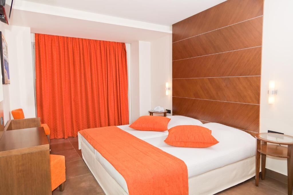 Une chambre d’hôtel avec un grand lit, une literie orange, une tête de lit en bois, un bureau avec une chaise et un rideau d’un orange vif.