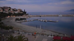 A coastal marina with boats docked, a promenade, and buildings in the background.