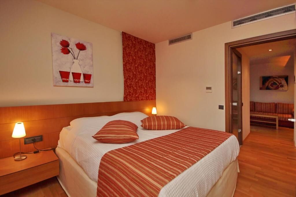 Un cuarto de hotel con una cama grande, dos lámparas de noche, un edredón blanco y rojo a rayas, una obra de arte floral roja en la pared, y piso de madera.