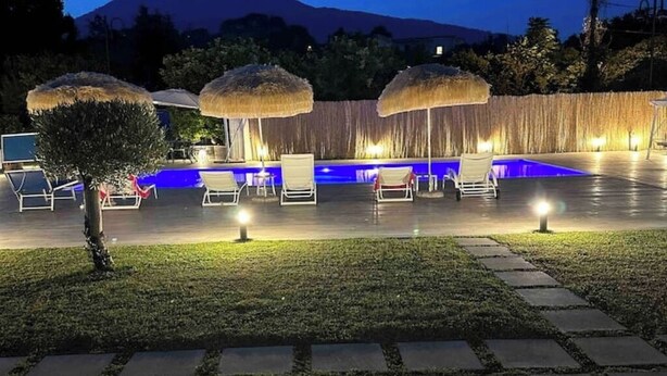 Area piscina con sedie a sdraio e ombrelloni, delimitata da un muretto e un piccolo albero.