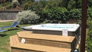 Una vasca idromassaggio in un giardino con un pontile in legno, circondata da vegetazione e una montagna sullo sfondo.