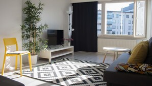 A modern living room with a sofa, a TV, a potted plant, a yellow chair, and a patterned rug. - Kotimaailma KotikolmioVesijk 3622 (Lahti)