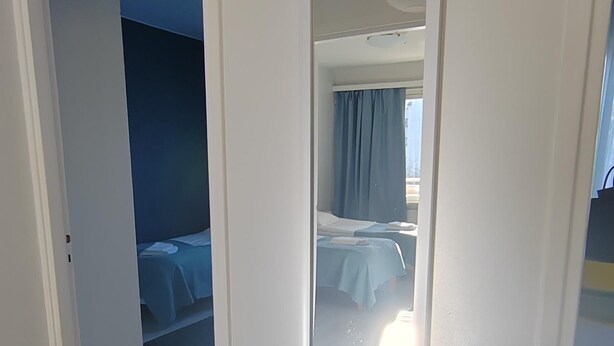 A hotel room with a white bench, a large mirror, and a door leading to a bedroom with a bed and a window with curtains. - Kotimaailma KotikolmioVesijk 3622 (Lahti)