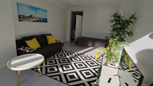 A modern living room with a black sofa, a white round table, and a patterned rug. There's a painting on the wall and a potted plant in the corner. - Kotimaailma KotikolmioVesijk 3622 (Lahti)