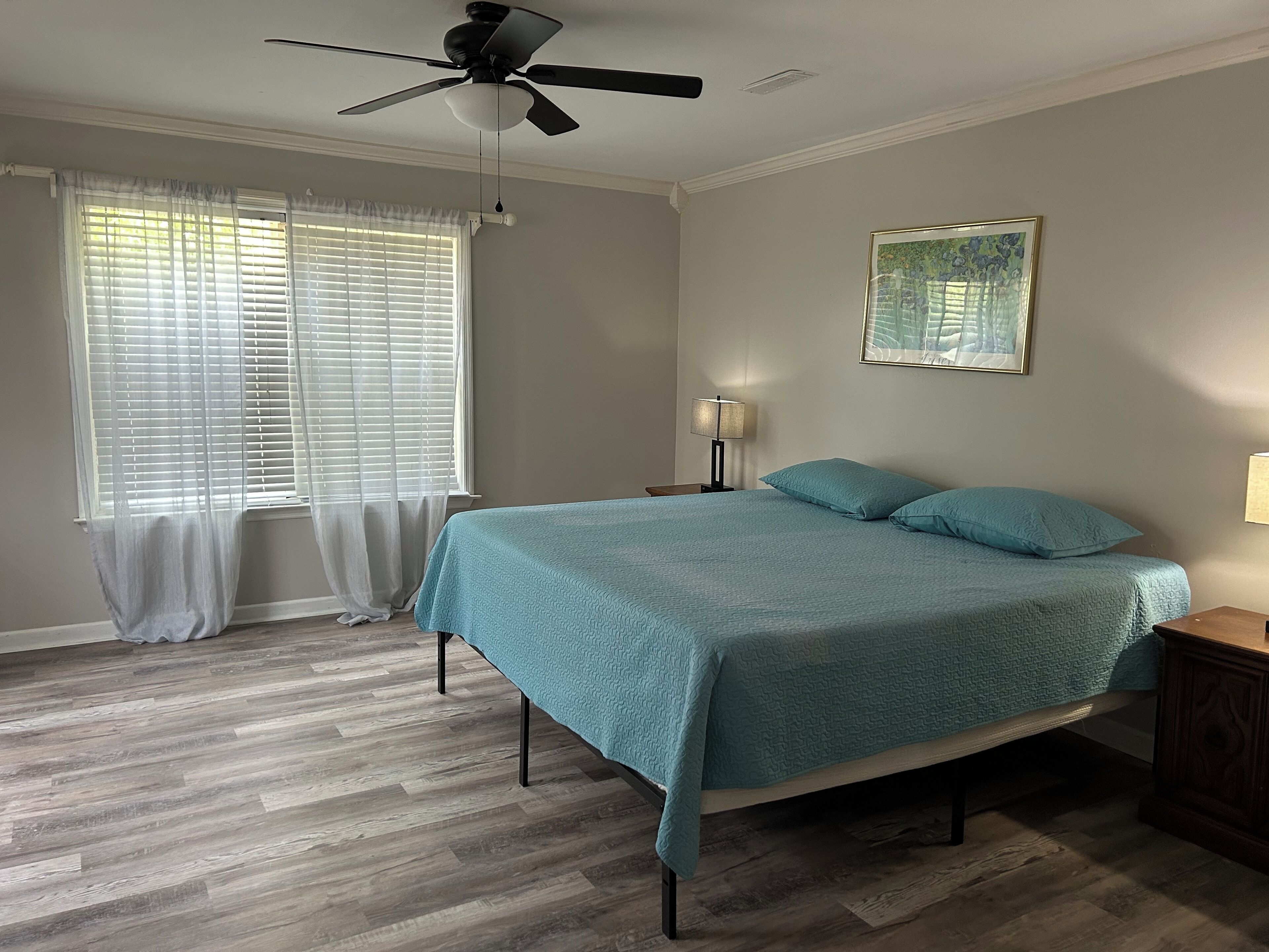 Ein Schlafzimmer mit einem Bett, Deckenventilator, Fenster mit Jalousien, Nachttisch und einer Wandlampe.