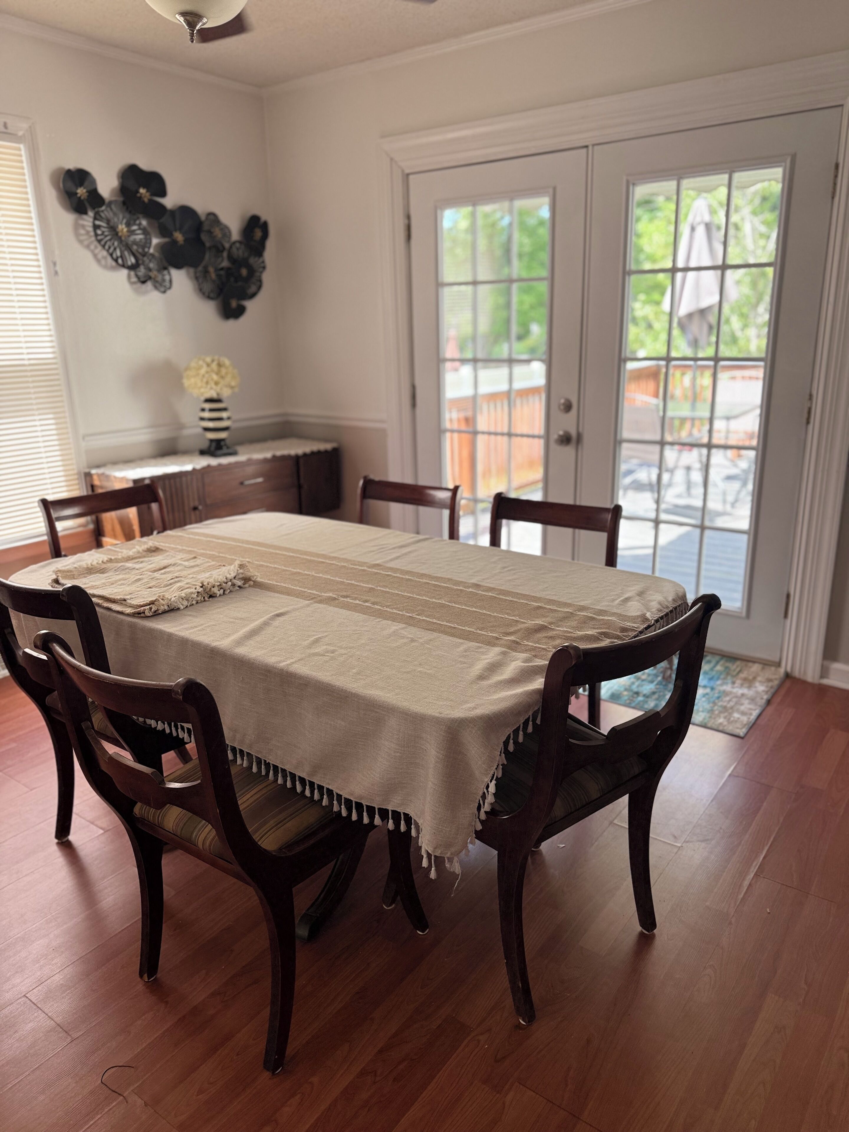 Une salle à manger avec une grande table, des chaises en bois foncé, un ventilateur de plafond et une vue sur un espace extérieur, accessible par une porte vitrée.